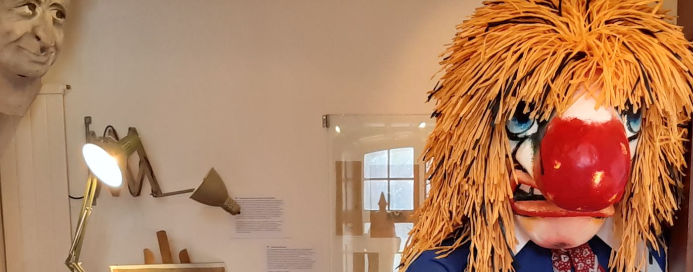 Rechts guckt eine Waggislarve mit riesiger roter Nase und wilden orangen Haaren aus dem Bild Dahinter sieht man in ein Larvenatelier