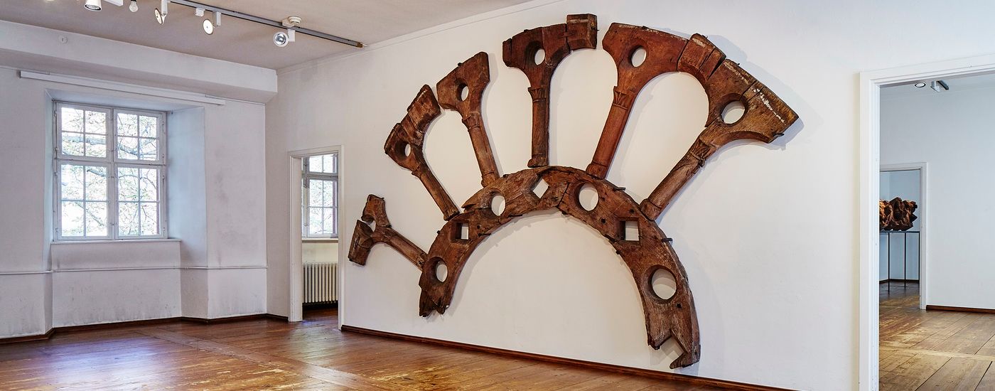 A large, fan-shaped wooden frame with circular openings hangs on a white museum wall