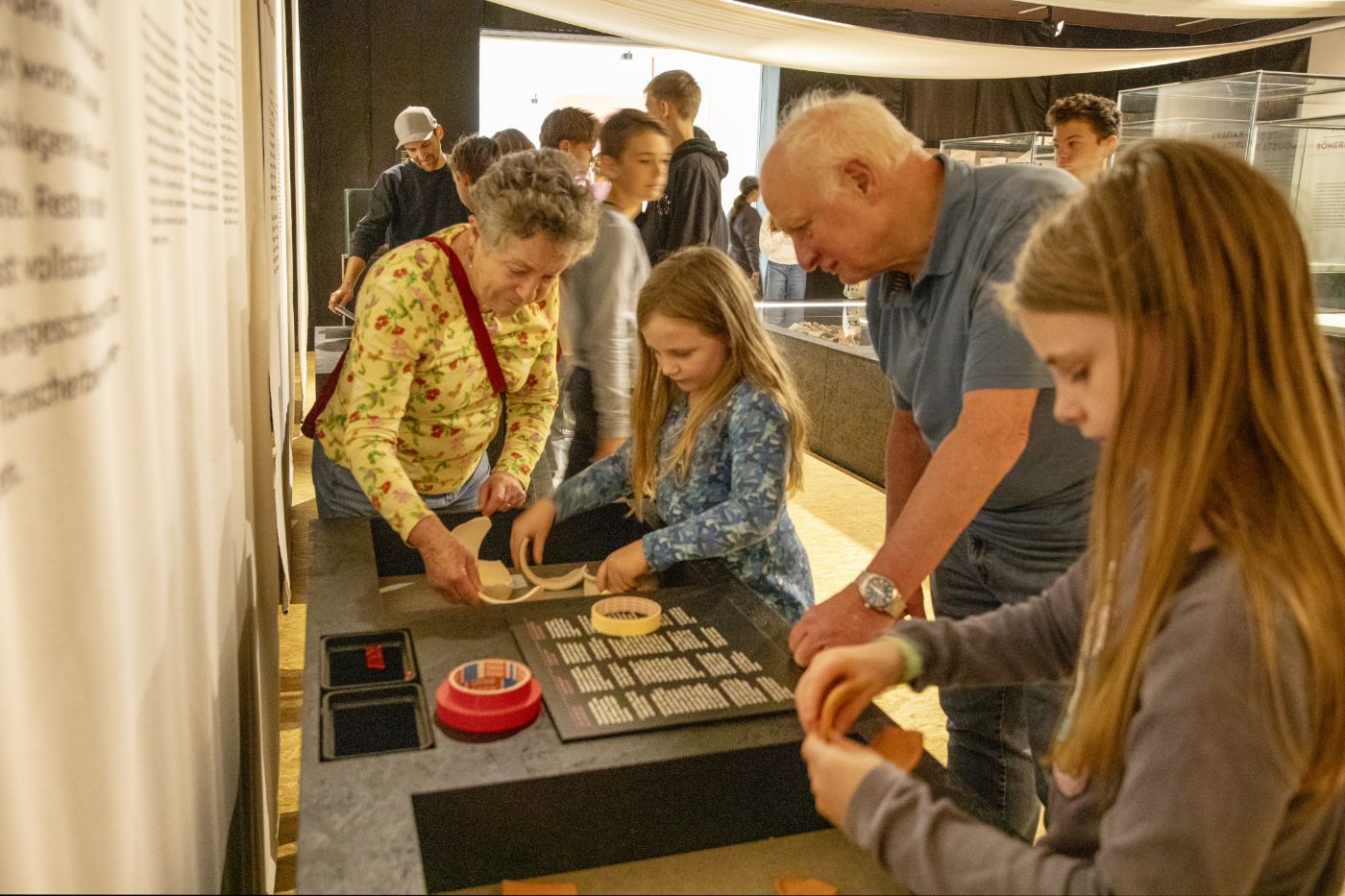 Eine interaktive Museumsszene mit mehreren Personen unterschiedlichen Alters, darunter ältere Erwachsene und Kinder. Eine ältere Frau und ein Mädchen stehen an einem Tisch und versuchen gemeinsam, Keramikscherben zu einem Gefäß zusammenzukleben. Ein älterer Mann und ein weiteres Mädchen beobachten die Aktivitäten konzentriert. Im Hintergrund sind weitere Besucher und Ausstellungsvitrinen zu sehen. Die Szene vermittelt eine Atmosphäre des Lernens und der Zusammenarbeit.