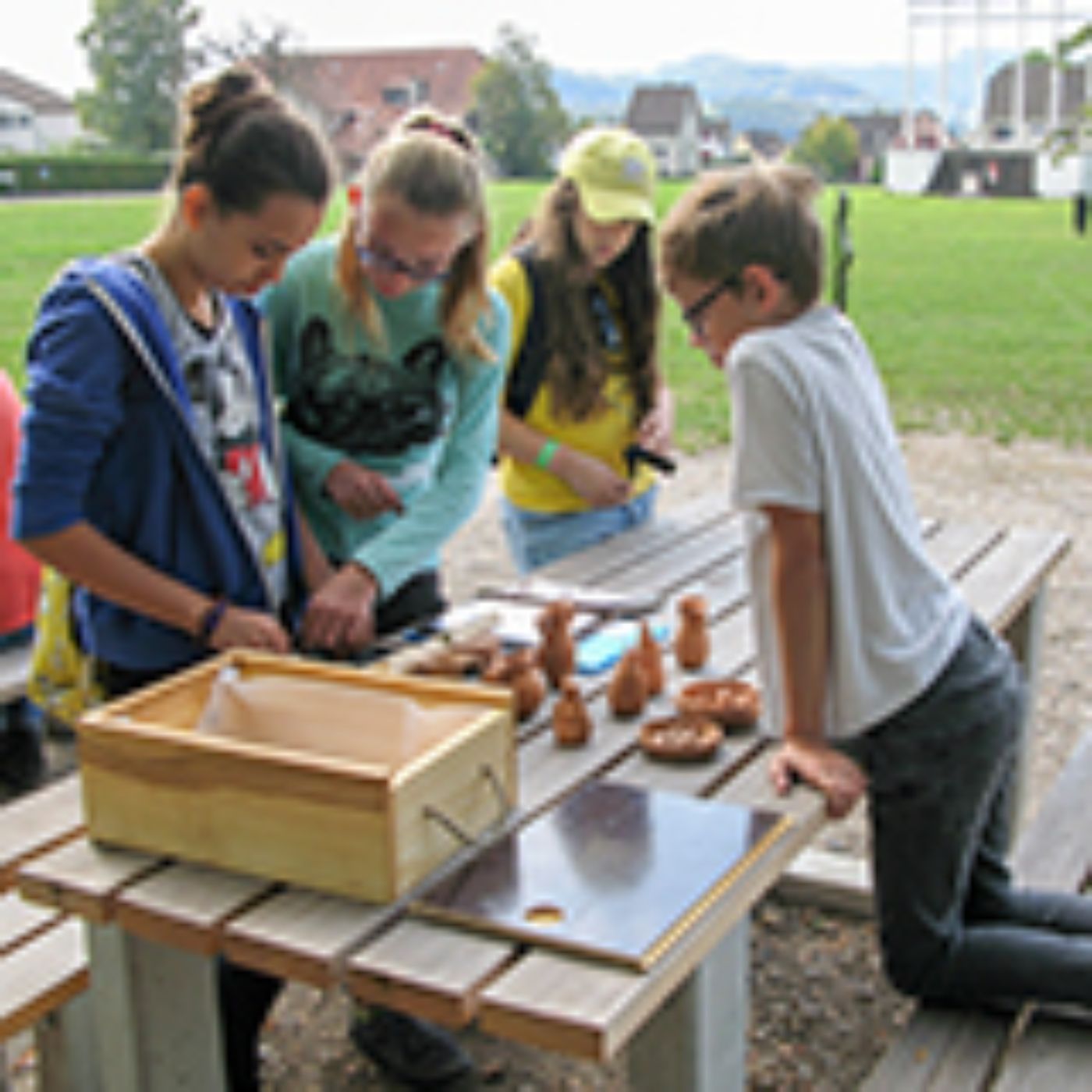 Auf einem Holztisch spielen vier Kinder im Freien mit kleinen römischen Spielfiguren