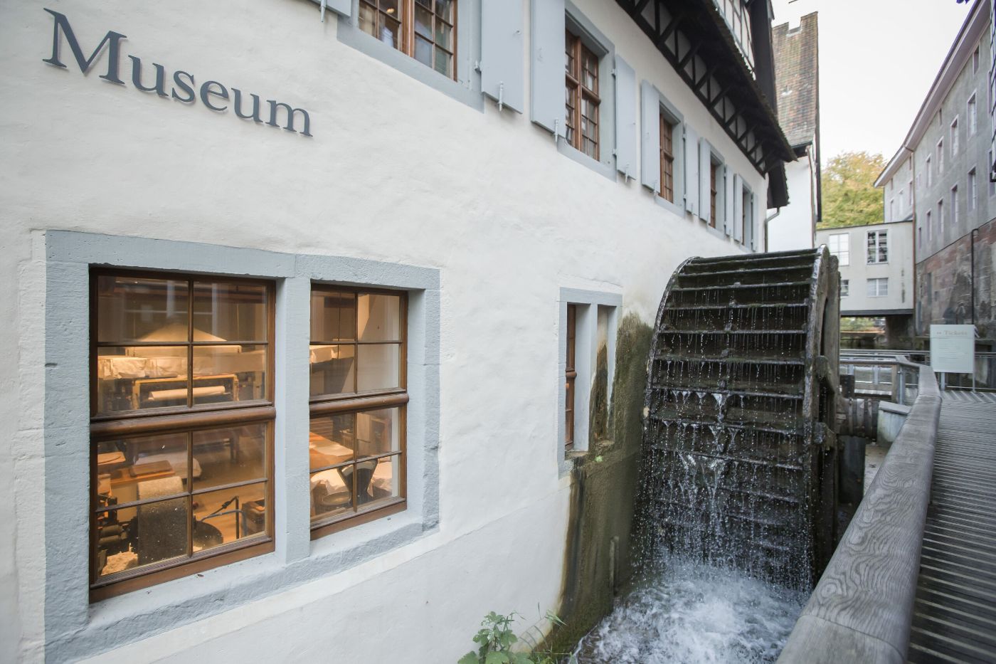 Museum mit Wasserrad und Fensterfront, sichtbare Inneneinrichtung und fließendes Wasser.