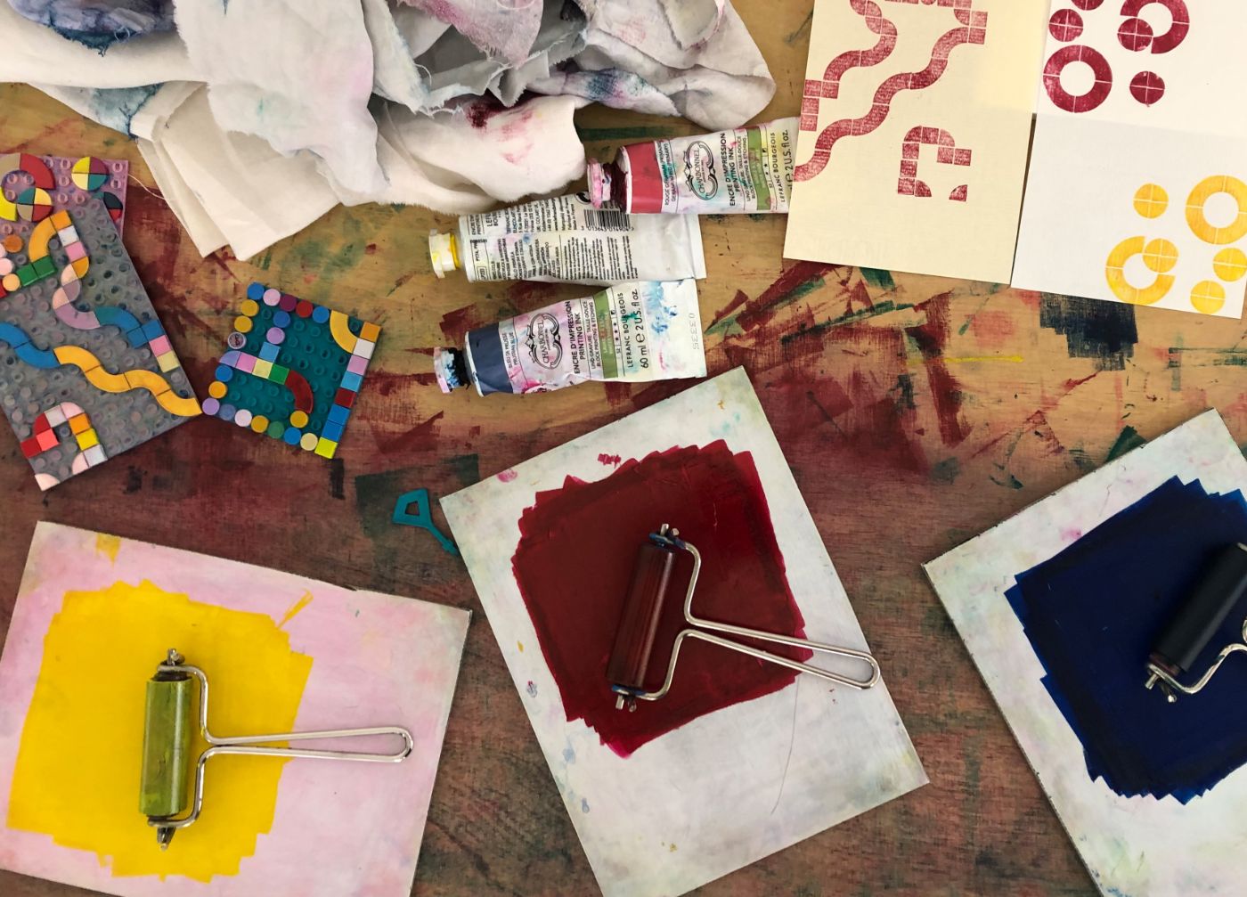 Bunte Farben mit kleinen Rollen und in Farbtuben, dazu werden die entstandenen Drucke ebenfalls gezeigt.Eine farbenfrohe, abstrakte Zeichnung mit verschiedenen Formen und Linien auf einem gelben Hintergrund.