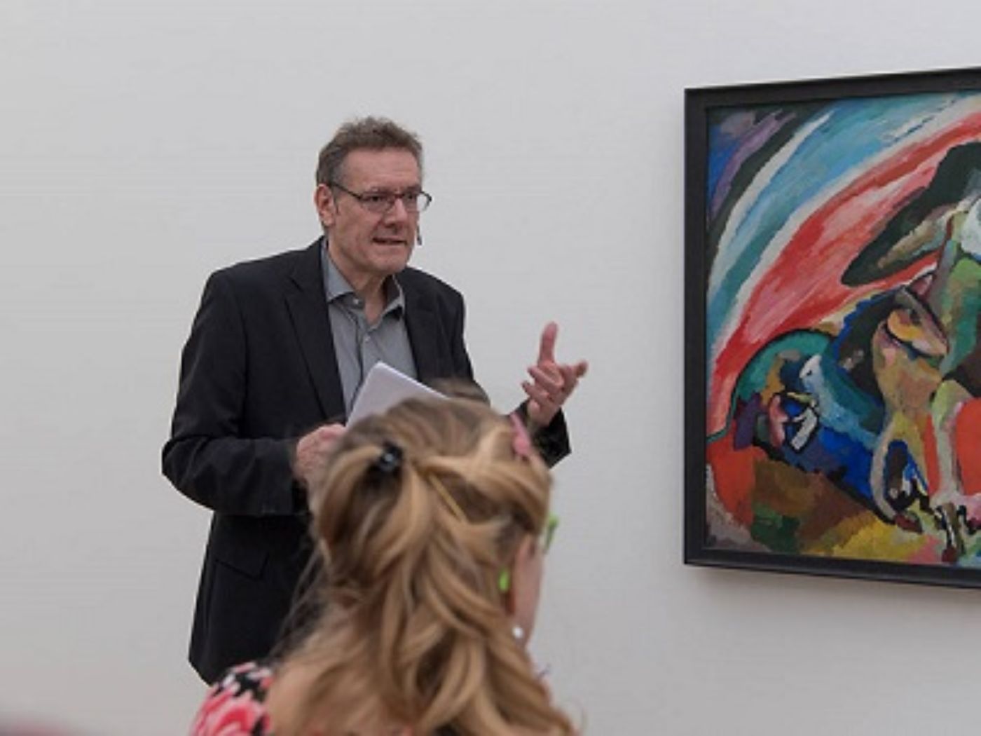 A man stands in front of a colorful painting, speaking to seated listeners