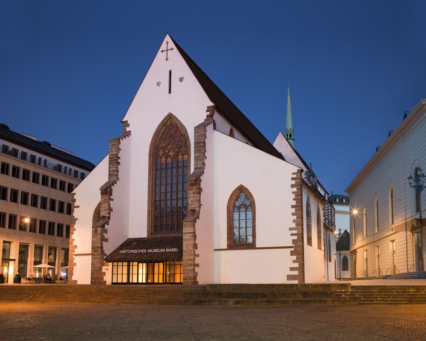 Historisches Gebäude mit großen gotischen Fenstern und beleuchteten Eingangsbereich am Abend.