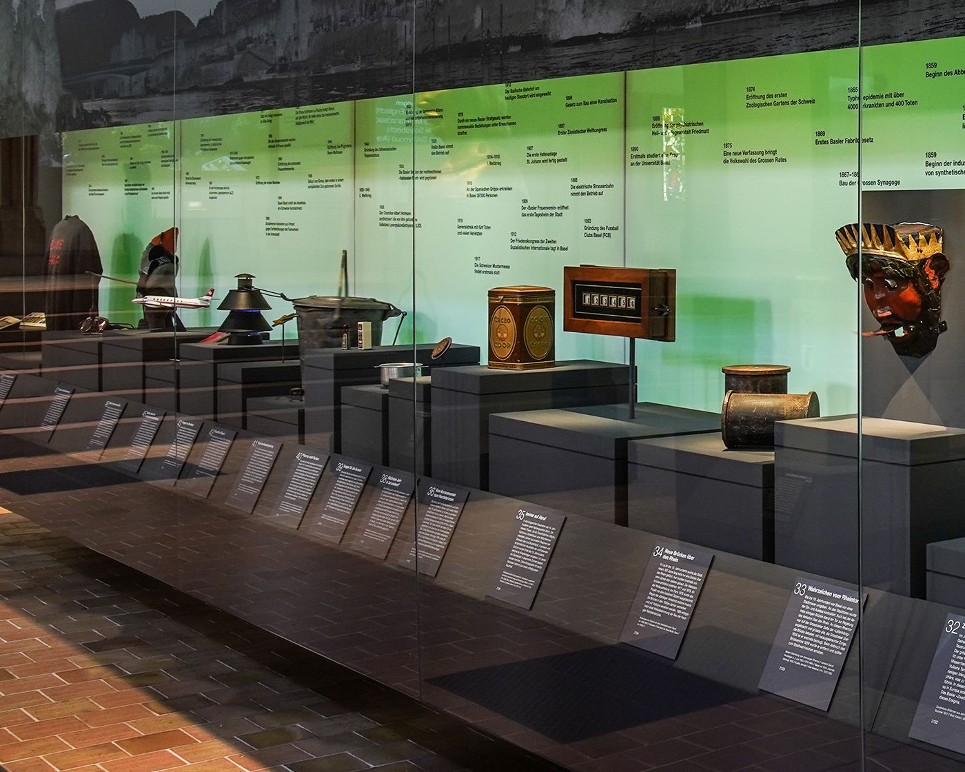 Auf einem grünen Hintergrund präsentieren Glasvitrinen historische Objekte mit Texttafeln davor