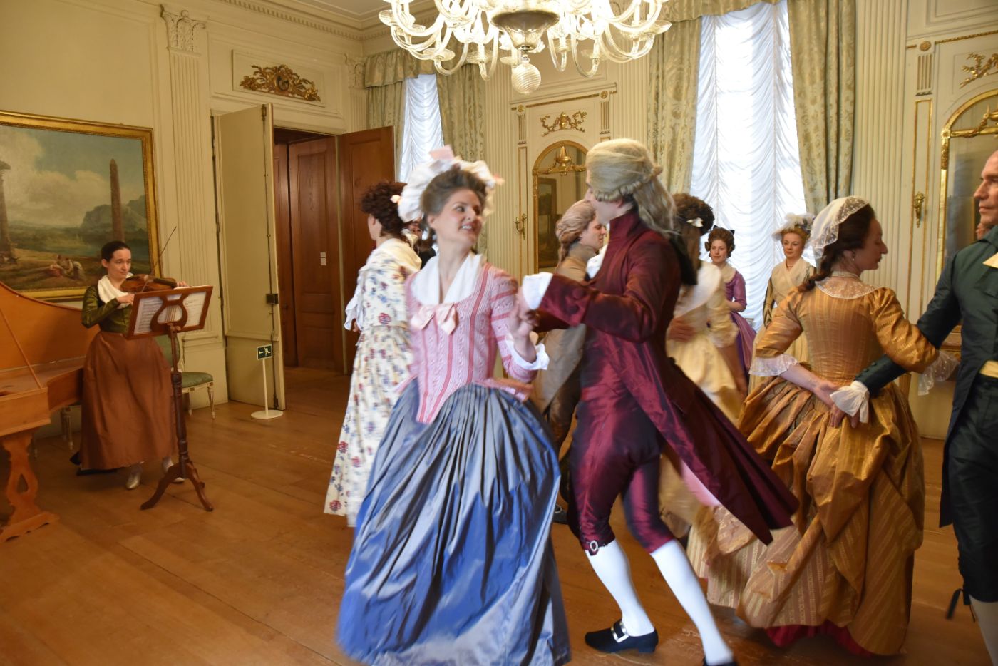 Eine Gruppe in eleganten Kostümen des 18. Jahrhunderts tanzt fröhlich in einem prunkvollen Saal