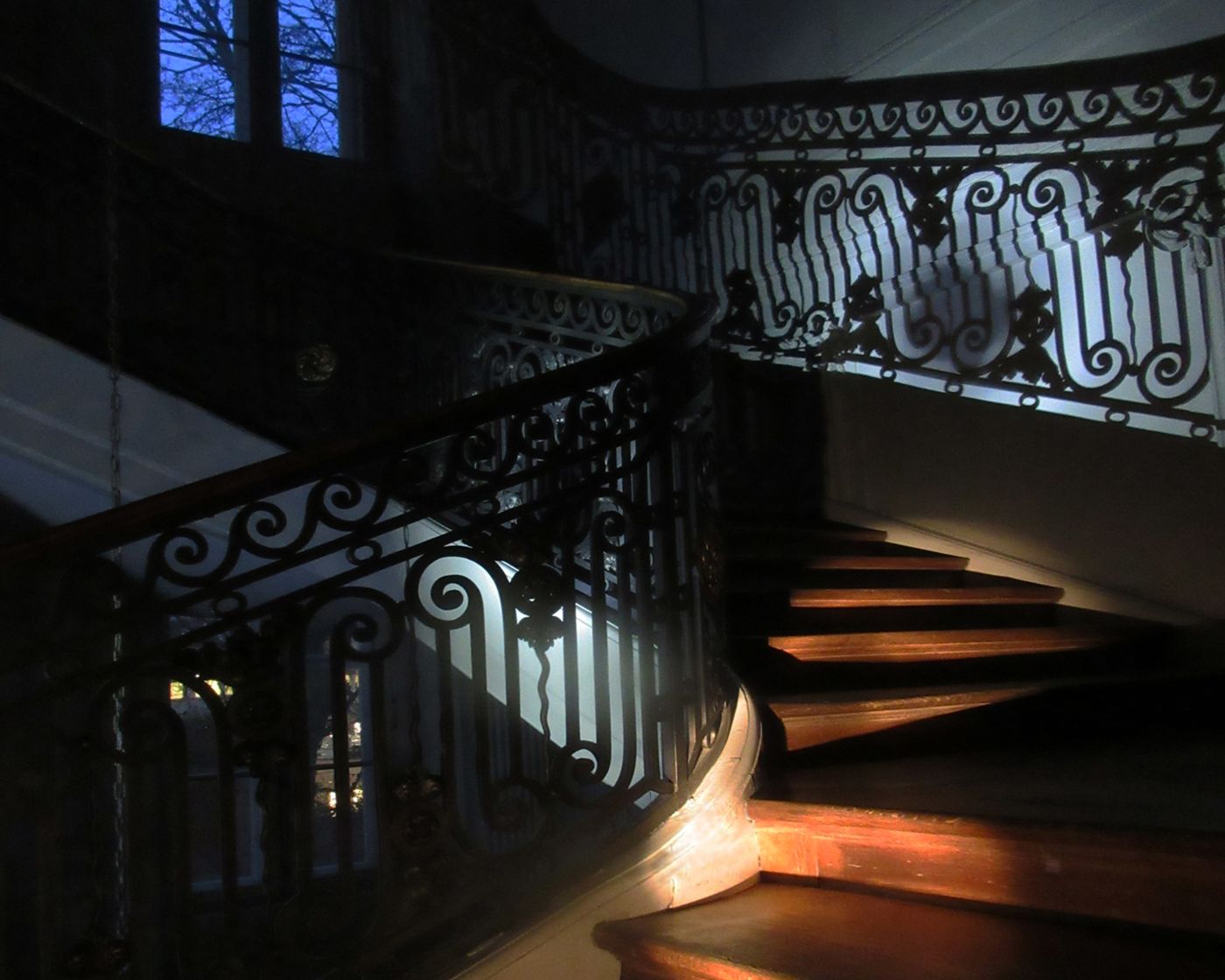 Eine geschwungene Holztreppe mit kunstvoll verziertem Eisen-Geländer im halb dunklen Licht