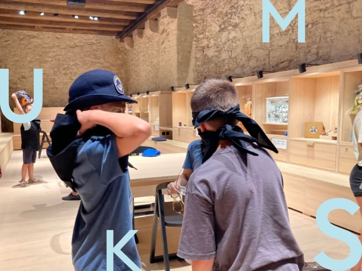 Auf dem Bild sieht man zwei Kinder mit verbundenen Augen, die in einem holzverkleideten Raum stehen