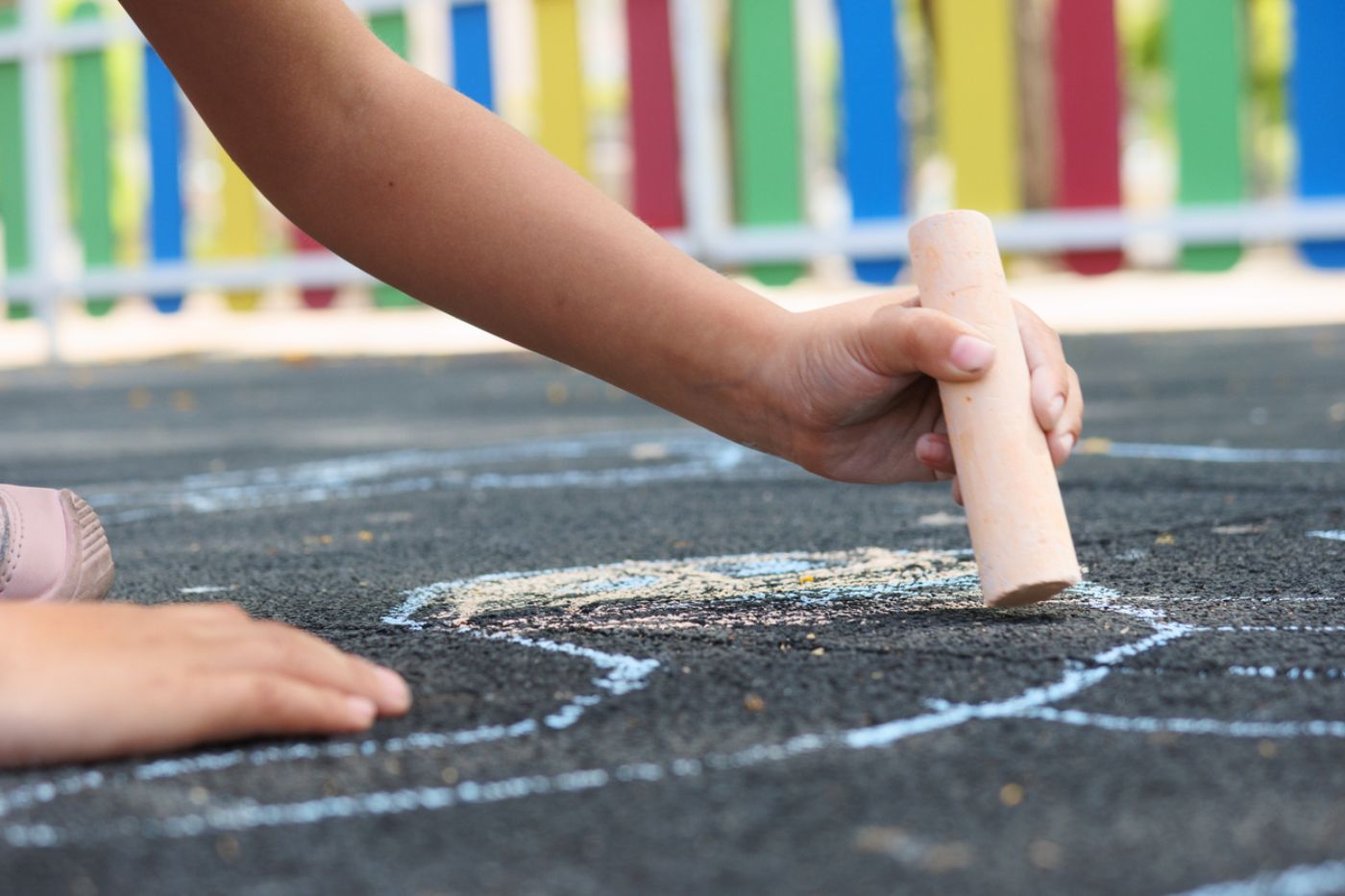 Ein Kind malt mit bunter Kreide auf Asphalt, der von einem bunten Zaun umgeben ist