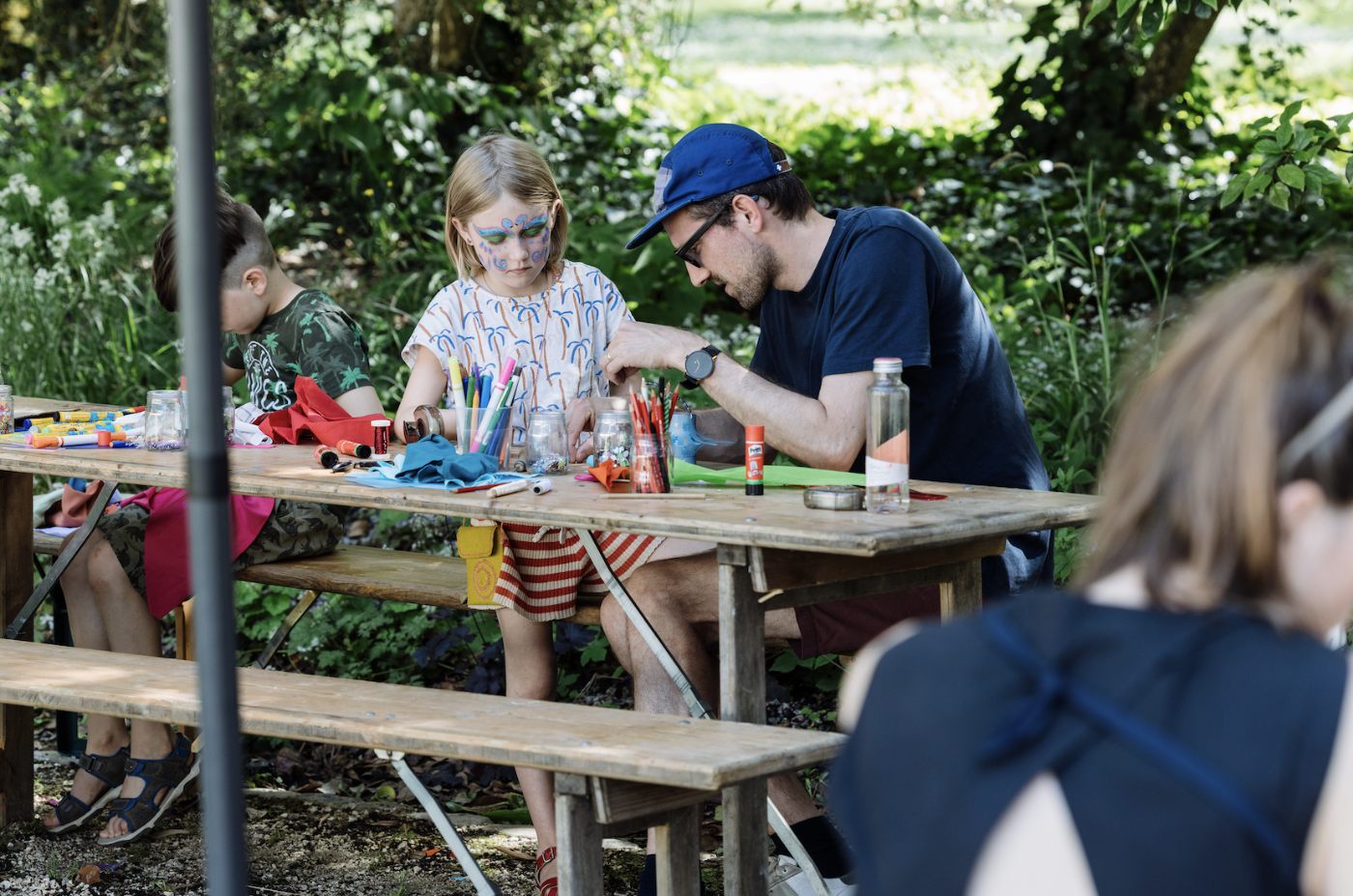 Deux enfants et un adulte bricolent avec des matériaux artistiques en plein air sur un banc en bois
