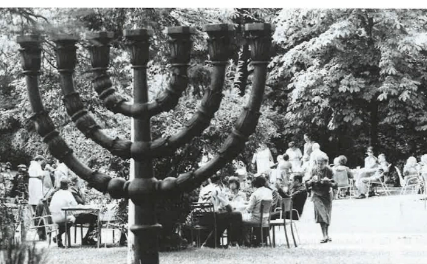 Eine grosse Menora steht im Freien; im Hintergrund versammeln sich Menschen unter Bäumen.