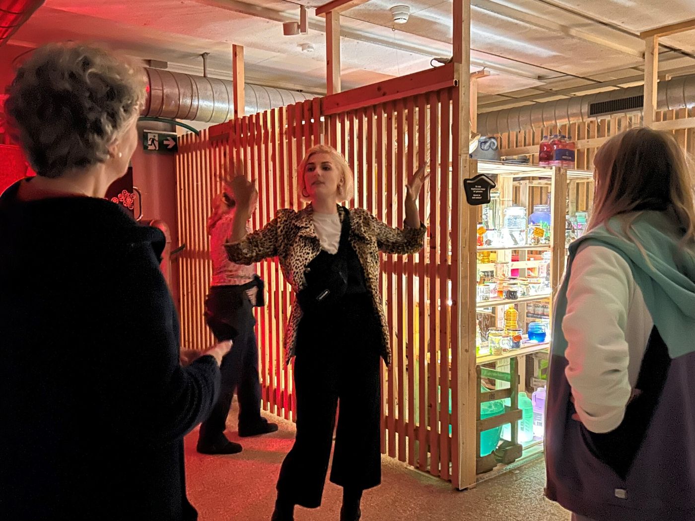 Eine Person in Leopardenjacke erklärt vor einer leuchtenden Holzwand in rot beleuchtetem Raum