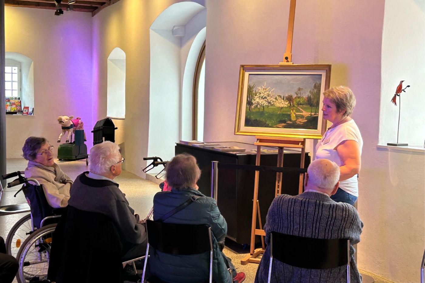 Eine Gruppe älterer Menschen betrachtet ein Gemälde in einem hellen Raum mit violetter Beleuchtung