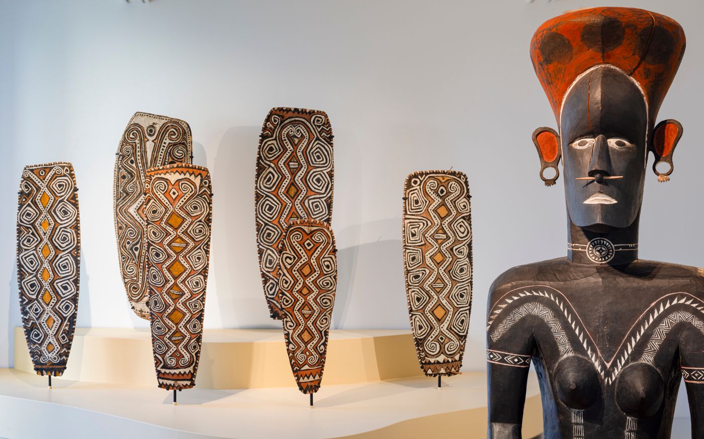 Figur aus braunem Holz mit roter Kopfbedeckung steht vor sechs Schildern, die reich verziert sind.