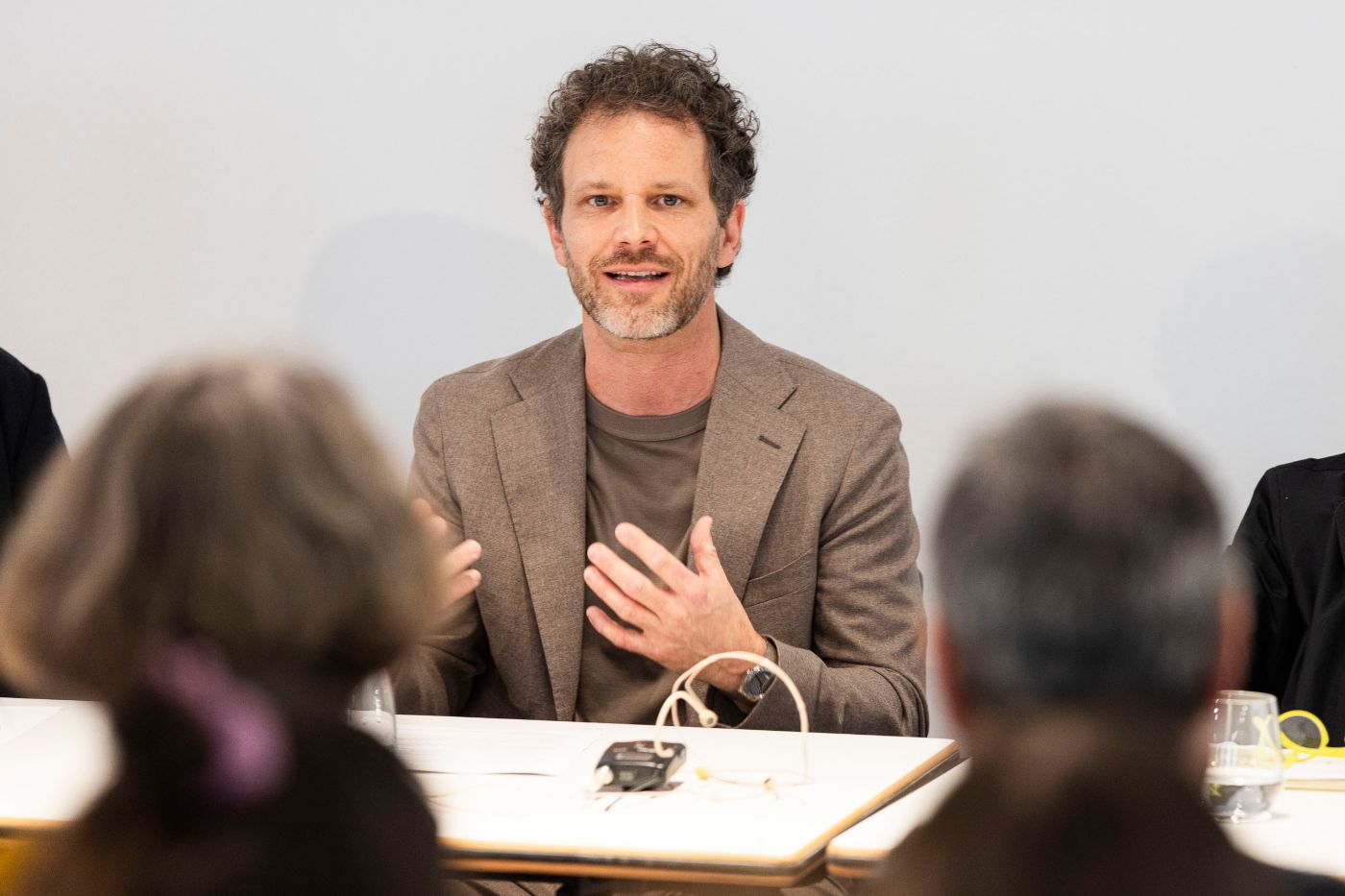 Ein Mann in braunem Anzug und braunem Pullover sitzt an einem Tisch und spricht. Vom Publikum sieht man nur zwei Köpfe von hinten.