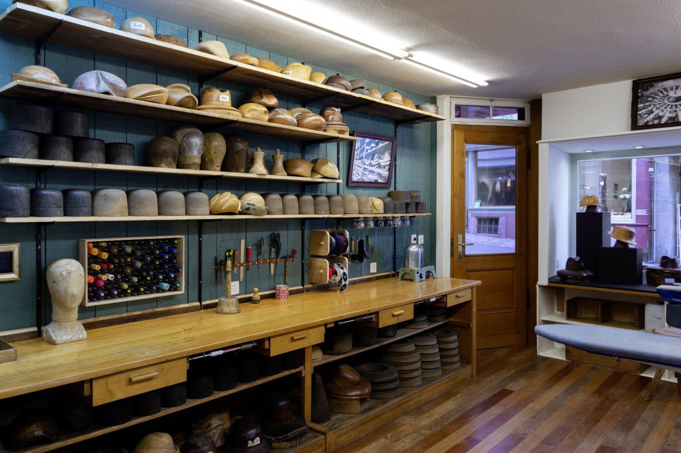 Geschäftsraum mit Tür in der Mitte, rechts davon ein Schaufenster, links davon hüfthohe Gestelle in Holz, auf denen Gipsköpfe stehen, darüber vier Regale mit Dingen drauf.