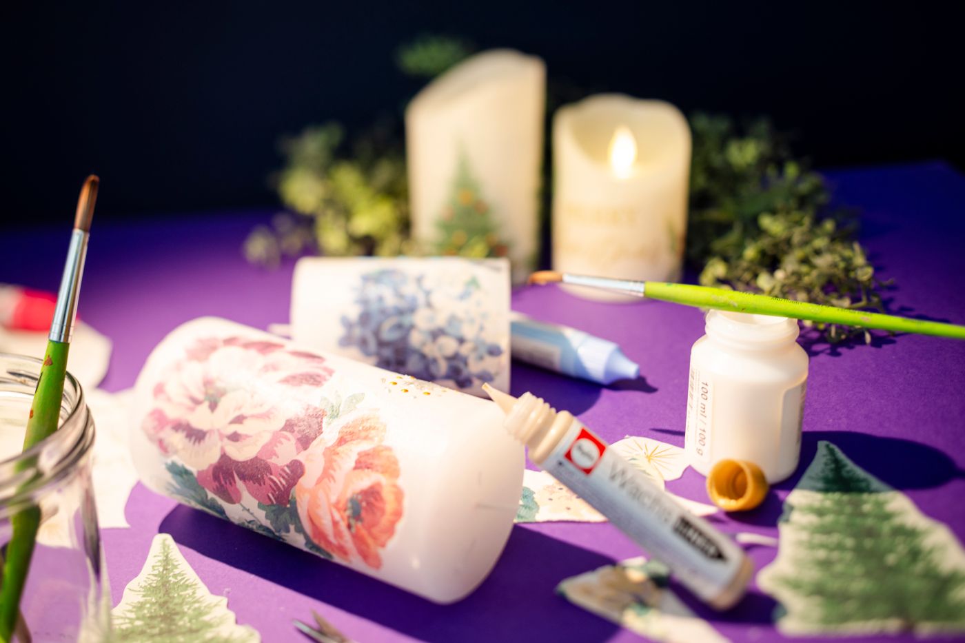 Auf einem violetten Tisch liegt eine weisse Kerze, die mit Blumenmotiven beklebt ist. Ein Leimstift ist an die Kerze angelehnt. Rundherum liegt anderes Bastelzubehör.