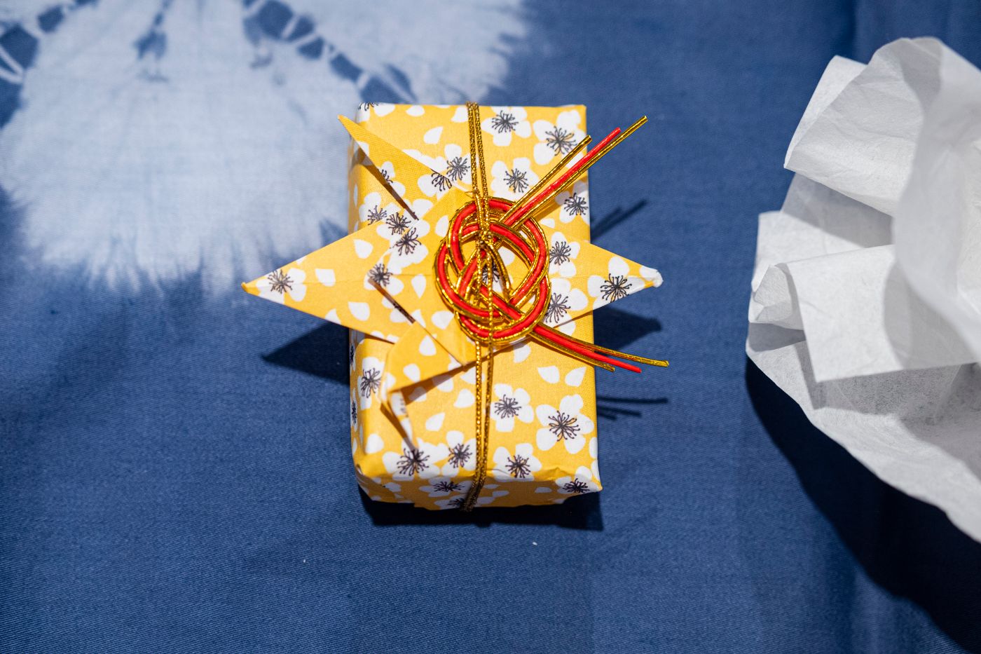 Auf einem blauen Tuch liegt ein rechteckiges Geschenk mit gelbem Papier und blau-weissen Blümchen. Der Geschenkbändel ist rot.