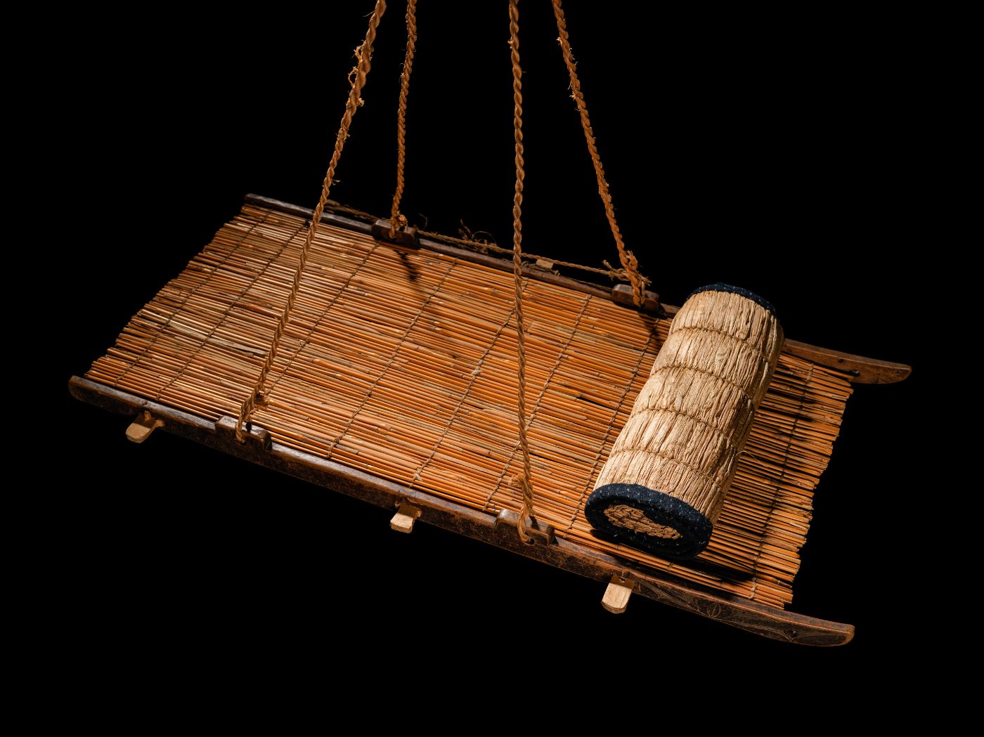 Bamboo and wood have been joined together to form a kind of board that hangs from four ropes; a mat is rolled up on top.