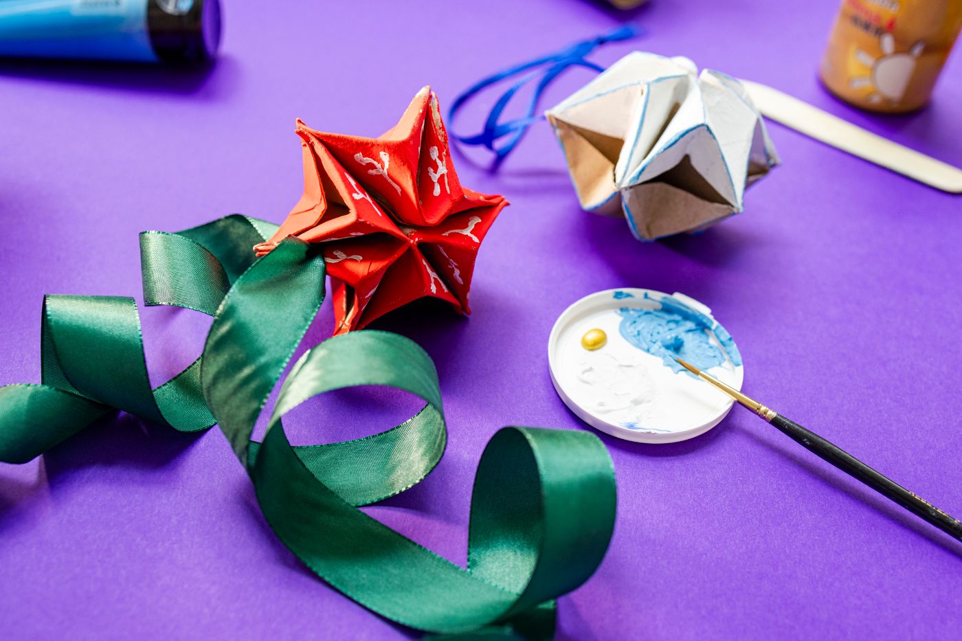 Auf einem violetten Tisch liegen ein roter und ein weisser aus Papier gebastelter Stern, daneben ein grünes Papierband sowie ein Pinsel in einem Deckelchen, in dem es auch blaue Farbtupfer gibt.