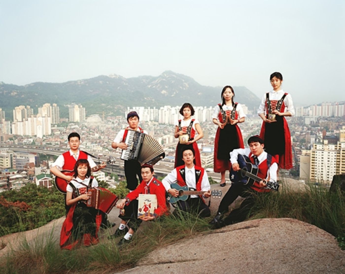 Auf einem Foto sind neun Personen in Trachten zu sehen, die Instrumente spielen, vor der Kulisse einer Grossstadt.