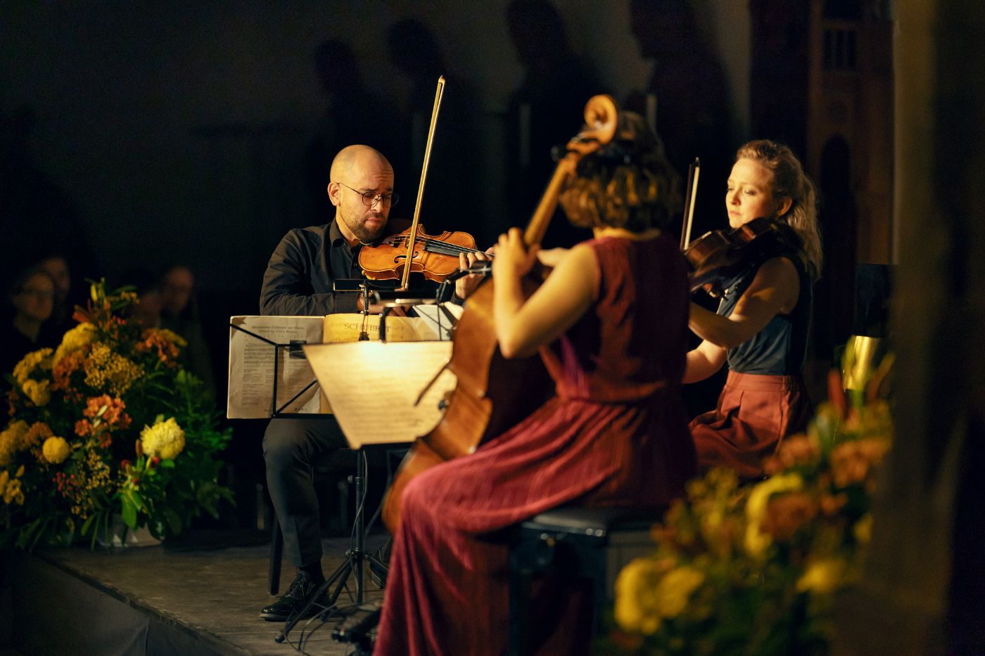 Ein Trio von Musikern spielt klassische Kammermusik, umgeben von Blumenarrangements.
