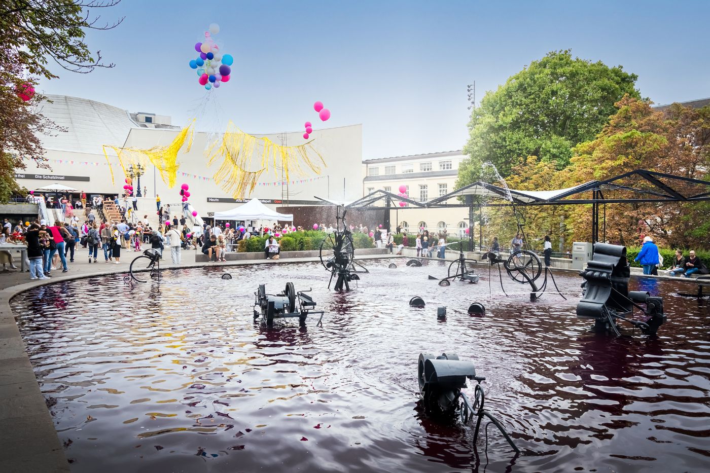 Ein lebhafter Platz mit Menschen, Luftballons und einem Springbrunnen mit Skulpturen im Wasser