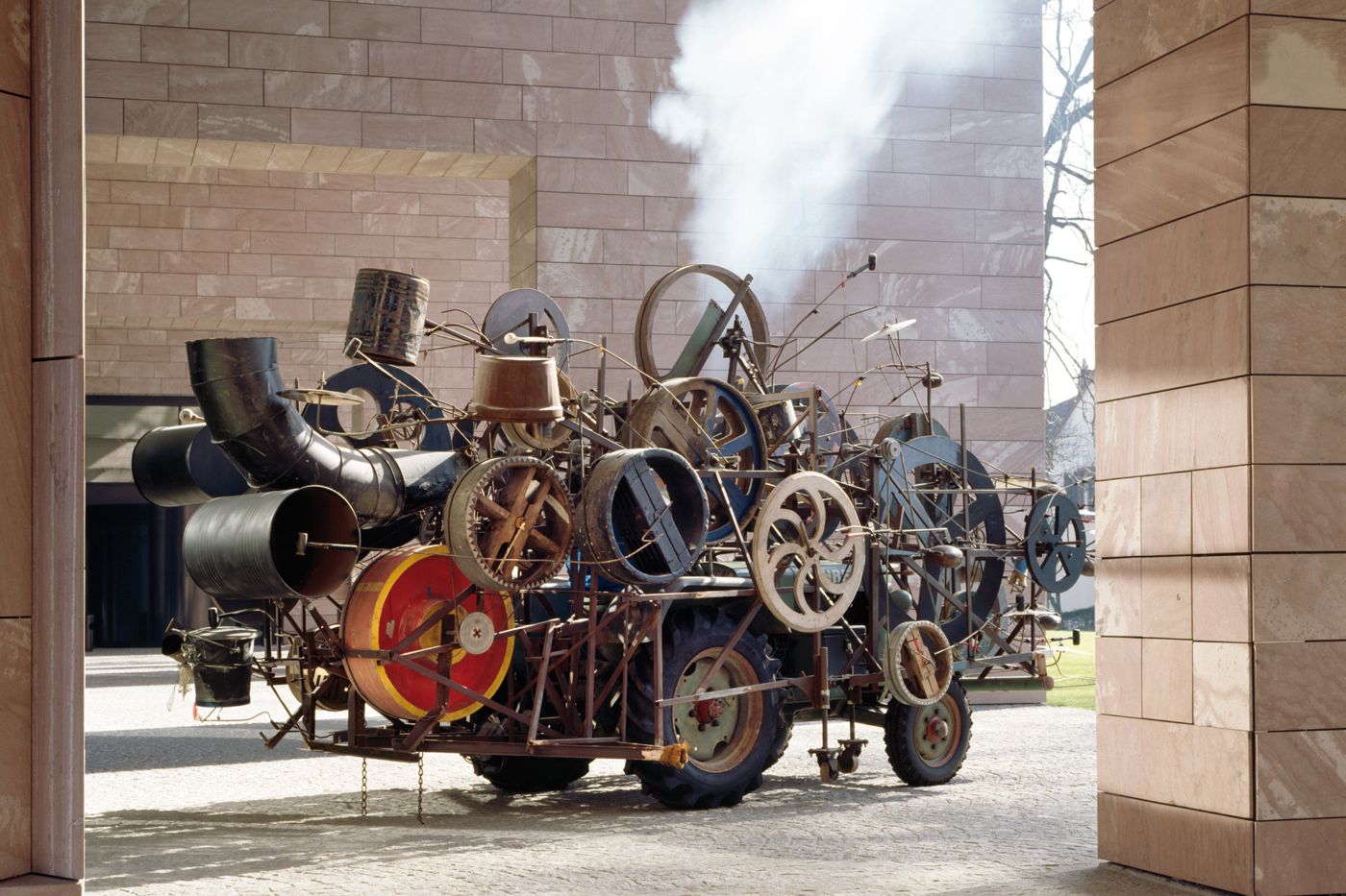A massive artwork of metal and wheels emits smoke and stands between two walls