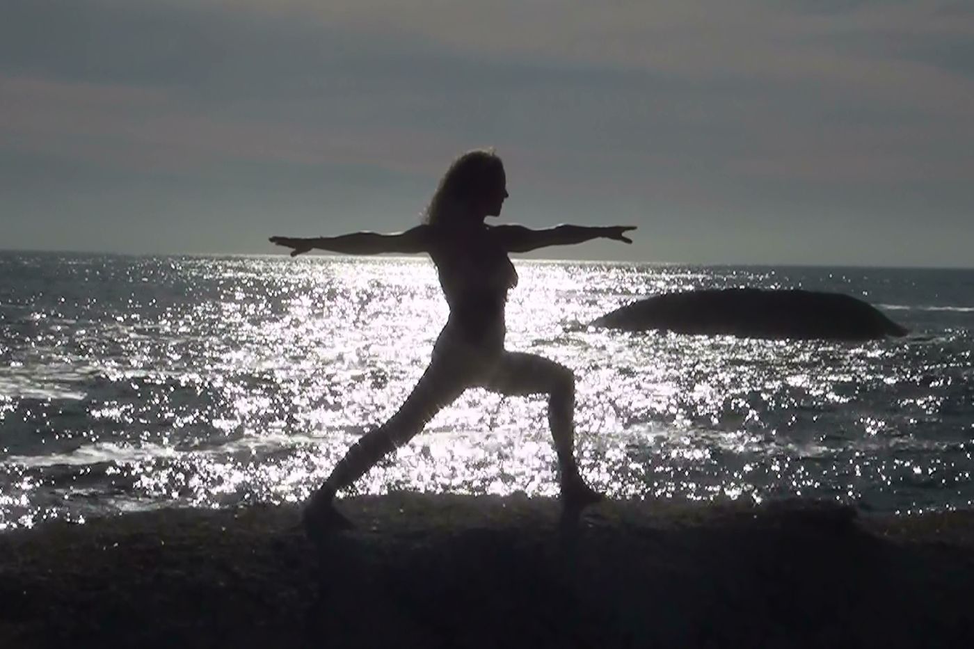 Eine Person führt eine Yoga-Pose am Meer aus, während die Sonne den Ozean funkeln lässt.