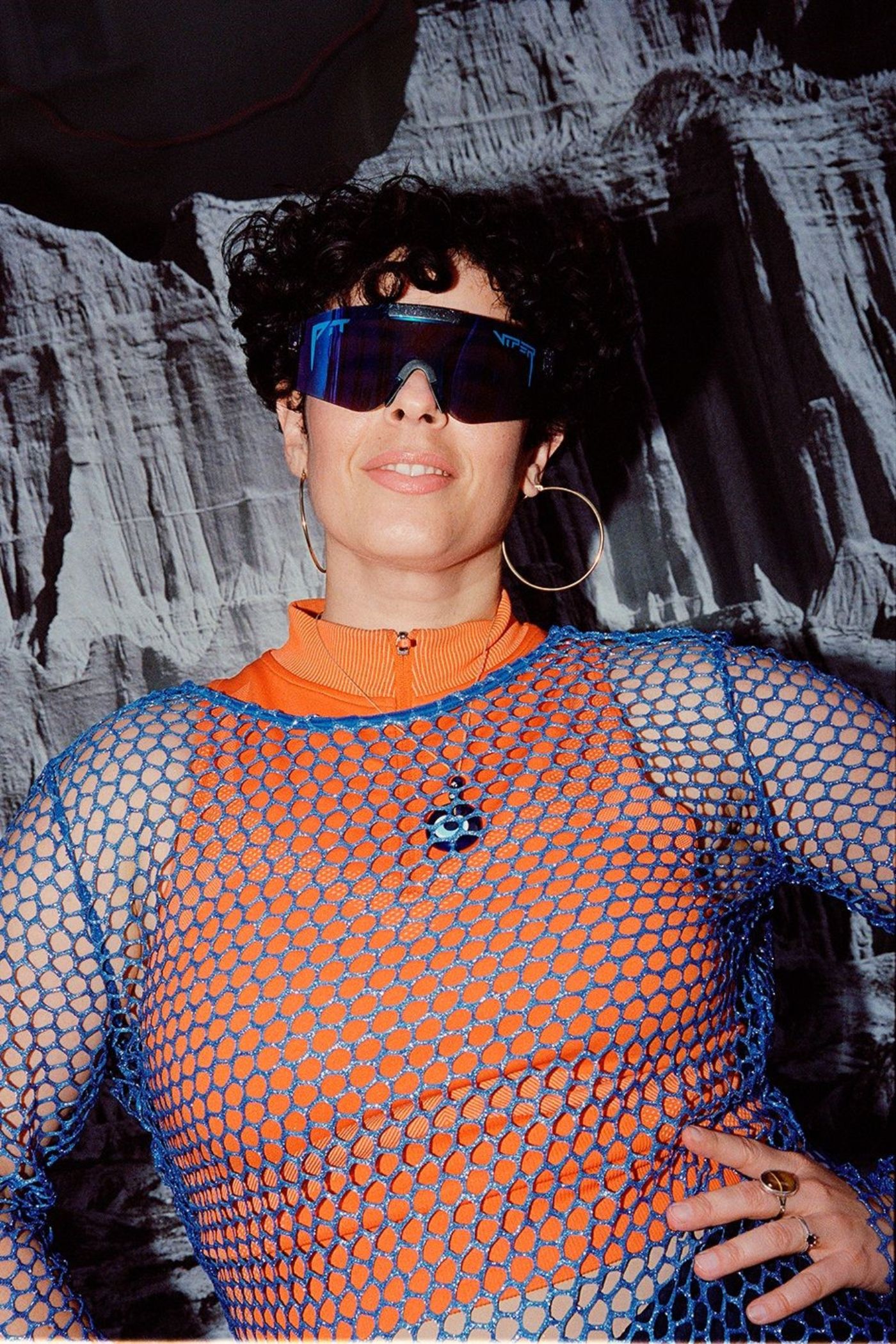 Person mit großer Sonnenbrille, blauem Netz-Oberteil und orangefarbenem Top vor grauem Hintergrund.
