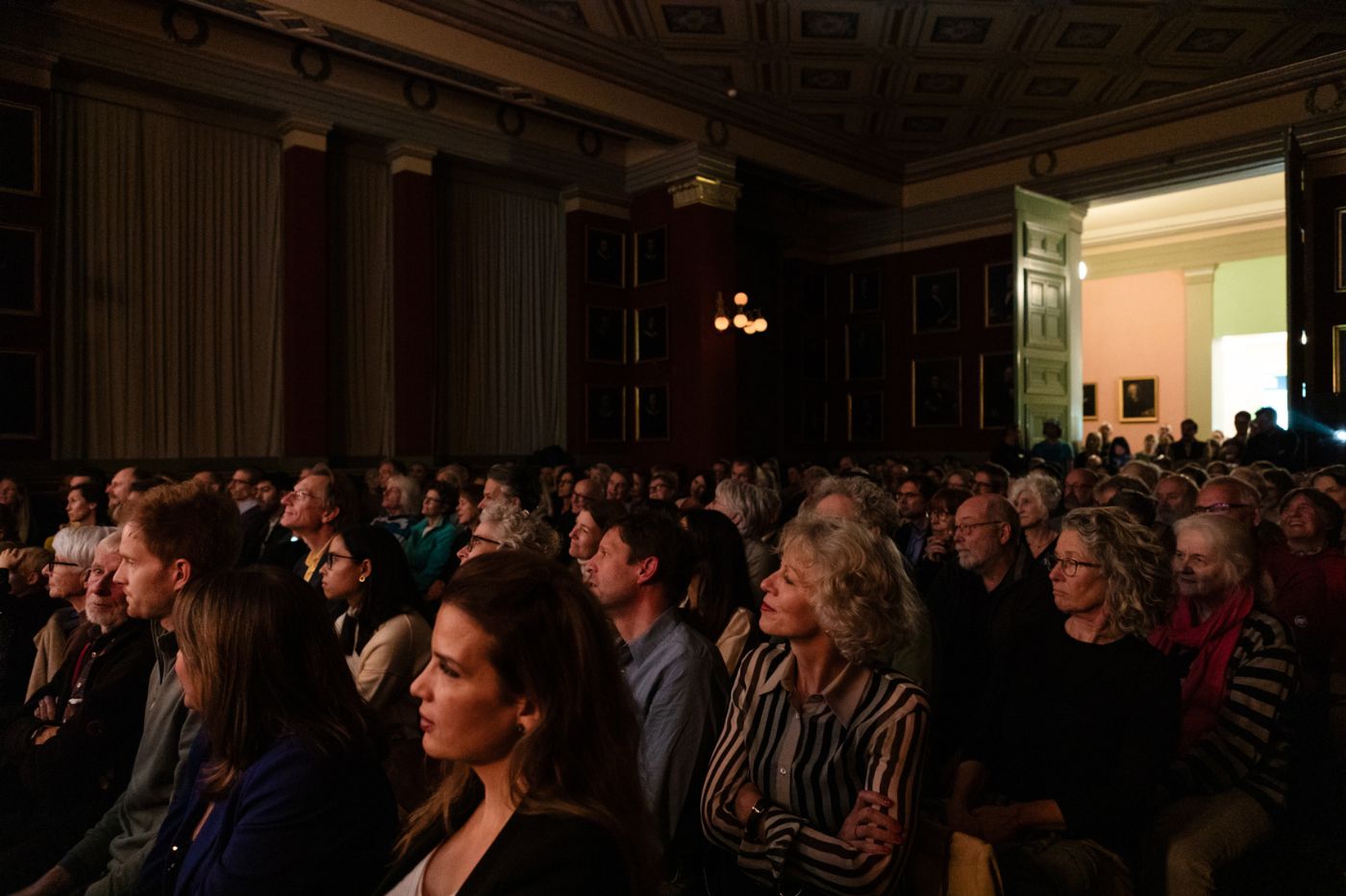 Eine grosse Menschenmenge sitzt in einem dunklen, eleganten Saal und schaut gespannt nach vorne