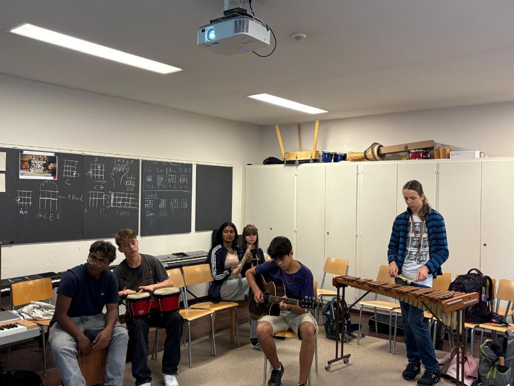 Mehrere junge Menschen sitzen und stehen in einem Schulzimmer mit Tafel und Kästen und spielen auf Instrumenten, auf einem Xylophon, auf einer Gitarre etc.