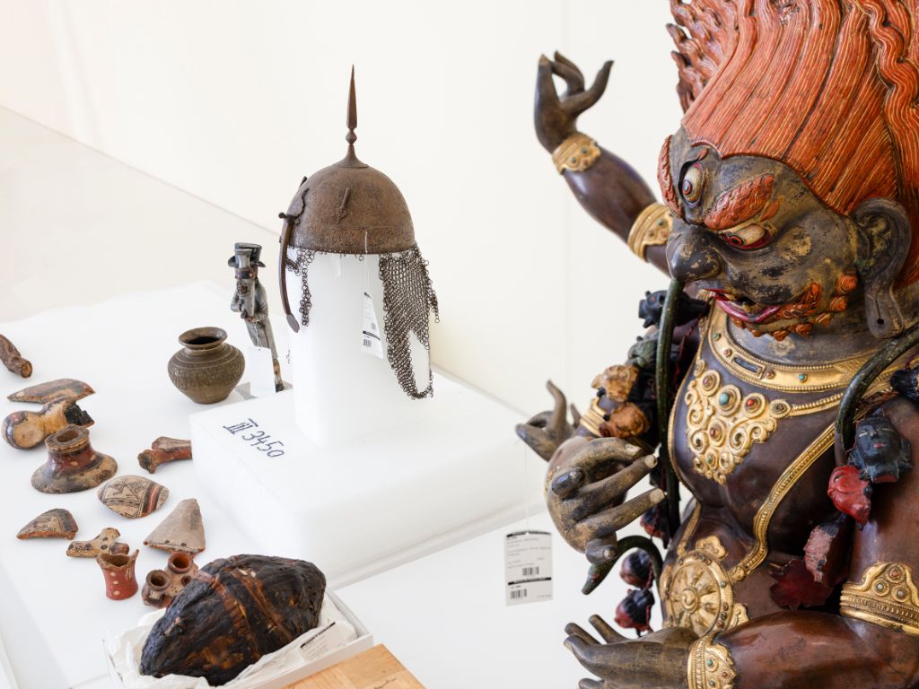 Auf einem weissen Tisch stehen und liegen etliche Dinge. Herausstechen eine Art brauner Helm auf einem Podest sowie eine Figur ganz rechts im Bild.
